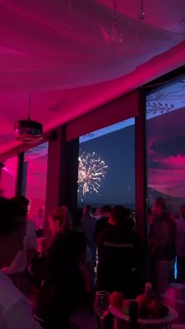 The video shows a group of people inside a dimly lit room with pink lighting, looking out a large window at a fireworks display in the night sky. The fireworks are bursts of orange and yellow light. Some people are holding drinks and talking, while others are focused on watching the fireworks. One person in the foreground is holding up a phone to record the display. In the room, there are tables with fruit and bottles on them. The ceiling is draped with white fabric.