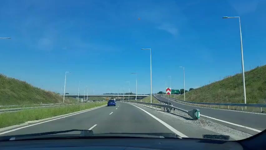 A POV shot from inside a vehicle driving on a highway on a clear, sunny day. The road has multiple lanes, with a white car in the left lane and a blue car further ahead in the right lane. Streetlights line the side of the highway, and a concrete overpass is visible in the distance. A guardrail runs along the right side of the road, next to a grassy embankment. A red and white chevron sign is placed on the embankment, indicating a curve. The sky is a bright, clear blue.