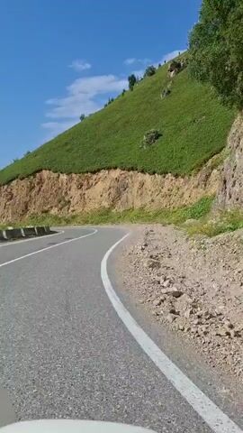 The video shows a view from inside a vehicle driving on a paved road. The road curves to the left, and on the right side of the road, there is a steep, grassy hillside. The hillside is cut by a sheer cliff face of tan-colored earth and rock, with some sparse vegetation. The sky is clear and blue with a few scattered white clouds. The vehicle's white bumper is visible in the bottom left corner of the frame. The scene appears to be a scenic mountain road.
