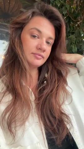 A woman with long, reddish-brown hair and fair skin is the main subject of this video. She is wearing a white button-up shirt over a black top. She looks directly at the camera, smiles, and winks. The background is slightly blurred, but it appears to be an outdoor seating area with green foliage and decorative lighting overhead.
