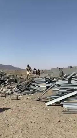 The video shows a group of men walking through a destroyed area. Debris is scattered on the ground, including metal, concrete blocks, and other materials. One man in a black outfit with a head covering walks toward the camera. Behind the debris are buildings, vehicles and mountains in the background. The sky is blue.