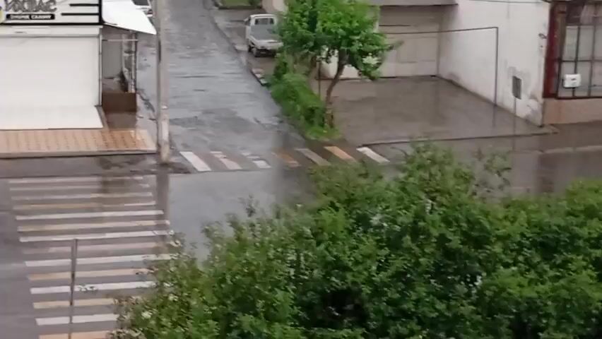 The video shows a street scene on a rainy day. The road and crosswalks are wet and reflective. There are buildings on the left side of the street, one with a white facade and a sign that reads "VOKSAS". A tree is growing in front of one of the buildings. Further down the street, a white van is parked. There are also trees and bushes visible on the right side of the street. The camera appears to be stationary and is looking down at the street from a slightly elevated position.