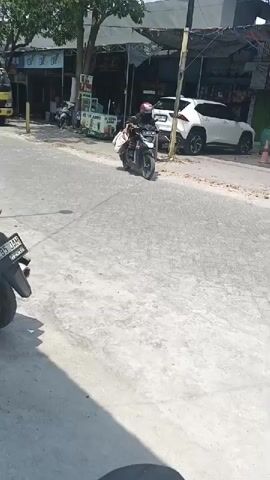 A person wearing a helmet and a dark outfit is riding a motorcycle. The motorcycle is carrying several packages, some of which are white and tied together. The motorcycle is traveling on a paved road in what appears to be an urban or suburban area. Buildings and other vehicles are visible in the background. The weather appears to be sunny.