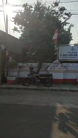 The video captures an outdoor scene, likely in a public space. The focus is on a building entrance, possibly a historical site or a cemetery, as indicated by the sign "Situs Makam R. Bagus Arya Wiralodra." The entrance is flanked by decorative brick pillars with red and white accents. Two Indonesian flags are visible, one on the left and one on the right side of the entrance, waving in the breeze. A motorcycle is parked on the sidewalk in front of the building, and another motorcycle is partially visible in the lower right corner of the frame. The sun is shining, casting shadows on the road, suggesting it is daytime. The overall impression is of a well-maintained historical marker or monument.