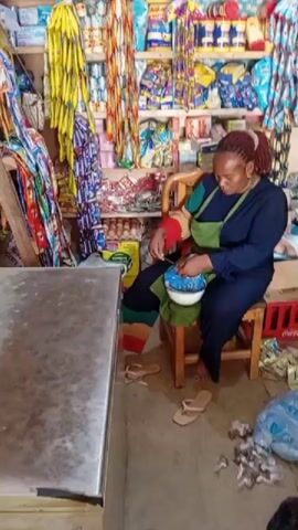 In a small, brightly lit convenience store in Gayawa, Nigeria, a woman with red/brown dreadlocks, wearing a dark blue top and an olive green apron, is seated on a wooden stool. She is actively mixing something in a white bowl with a utensil.

The shop is densely packed with various products. Shelves along the back and right walls are fully stocked with packaged goods, including food items, drinks, and what appear to be toiletries, with many items also hanging from the ceiling. On the floor near the woman, two pairs of flip-flops are visible, along with a blue plastic bag containing small white objects and some scattered debris. A red Coca-Cola crate and a carton of eggs are also present. To the left, a metal counter or table is visible. The woman appears focused on her task. The blue plastic bag has "JEAN" printed on it.
