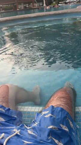 A person wearing blue swim trunks with a white shark pattern sits in a swimming pool, with their legs submerged in the water. The pool has small blue tiles and water that gently ripples, reflecting the surrounding light. In the background, there are other people in the pool, as well as lounge chairs and a covered structure. The video is taken from a first-person perspective.