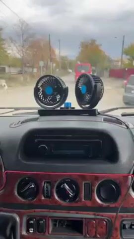 A view from the dashboard of a vehicle traveling on a road in a rural-esque area. The dashboard is dark with what appears to be wood grain accents. A pink light strip runs along the base of the dashboard, below various dials and knobs. On top of the dashboard, there is a stereo system and a small stand holding two mini fans with blue centers. The fans are pointed forward. Through the windshield, a road stretches ahead with buildings and trees on either side. A red van is driving away in the distance. The sky is overcast. The car is moving forward on the road. There is no discernable human presence in the immediate shot aside from the implied driver.