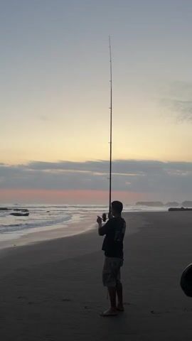 A man stands on a sandy beach at twilight, fishing with a long rod. He faces the ocean, his back to the viewer. The sky is a gradient of pale yellow, pink, and light blue, with some clouds. Waves gently crash on the shore, and rocky outcrops are visible in the distance to the right. The man wears a dark shirt and light-colored shorts. He is barefoot.
