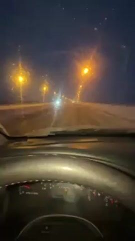 The video, shot from the driver's seat of a car at night, shows a snowy road illuminated by streetlights. The dashboard is visible, displaying gauges and indicators. The windshield is partially covered with snow, with some areas cleared by the wipers. Other cars' headlights can be seen in the distance.