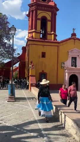 The video shows a sunny day in front of a yellow and red church with a tall bell tower. People are walking around the church, including a woman in a white hat and blue skirt who is walking towards the camera. A couple walks past a large stone cross in the foreground. The camera pans up to show the blue sky with a few clouds. The sound of church bells can be heard.