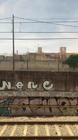 A train is moving along train tracks. On the right side of the tracks, there is a concrete wall covered in graffiti. In the background, there are buildings, trees, and power lines. The sky is partly cloudy. The graffiti is colorful and consists of various tags and designs. Some of the graffiti is obscured by shadows or vegetation. A few cars are visible in the distance. The train appears to be moving through an urban or suburban area.