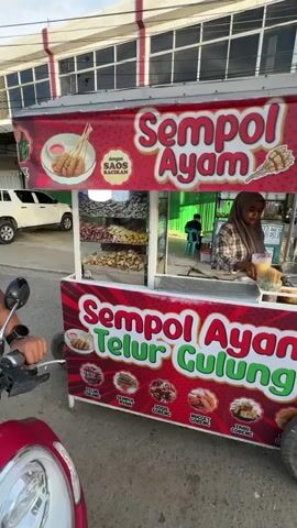 The video shows a food cart with red and white branding that says "Sempul Ayam" and "Sempul Ayam Telur Gulung". There is a woman wearing a hijab behind the counter. She appears to be preparing food. Various snacks are displayed in trays on the cart. The cart is on a street, with a building in the background. A motorcycle is partially visible in the foreground.
