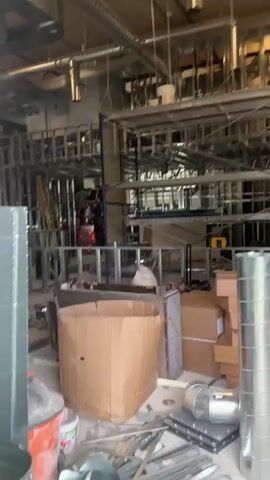 The video shows an indoor construction site with metal framing and exposed ventilation systems. Several workers are present, some on a scissor lift, appearing to be engaged in construction tasks such as framing or installing materials. Various construction materials, including cardboard boxes, metal pipes, and tools, are scattered around the site. The environment suggests an ongoing renovation or new construction project.