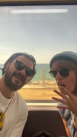 A man and a woman are sitting next to each other in what appears to be a train car. They are looking into the camera, smiling and singing along to music playing. The man is on the left side of the frame, has a beard, and is wearing sunglasses and a white t-shirt with a necklace. The woman is on the right side of the frame. She's also wearing sunglasses and a hat. Through the train window, there is a clear blue sky, ocean water, and a sandy beach with cyclists.