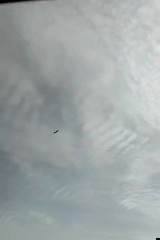 A drone is shown flying in the sky. The drone is black and is silhouetted against a cloudy sky. The clouds are white and fluffy, and they appear to be in layers. The drone is flying at a high altitude, and it is difficult to make out its features. The drone is moving from left to right across the screen. The video is shot from below, looking up at the drone.