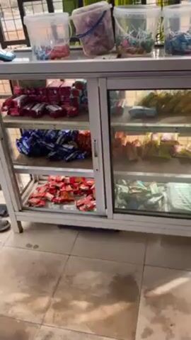 The video shows a display case filled with packaged food items in what appears to be a store or shop. The case has a white frame and sliding glass doors. The shelves are stocked with various types of packaged snacks and candies. Above the display case are several clear plastic buckets, also containing different types of sweets or candies. The floor is tiled. A pair of blue sandals is visible near the bottom left corner of the frame. The scene is brightly lit. There is Arabic-sounding background audio.