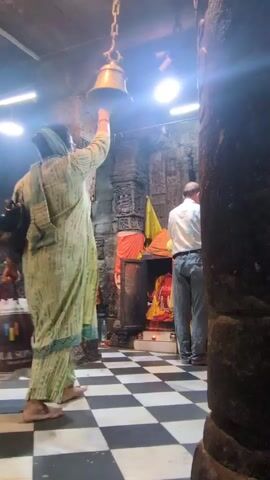 A woman in a light green patterned salwar kameez and headscarf rings a brass bell hanging from a chain above her head. She is barefoot. To her right, another person in a white shirt and grey pants stands facing away from the camera. The floor is a black and white checkered pattern. The background appears to be a temple, with stone pillars and deities visible. The lighting is dim, with bright spotlights overhead. A bag is slung over the woman's left shoulder. The video is shot from a low angle.