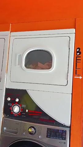 This is a close-up, vertical shot of a stacked laundry unit. The top portion is a white dryer with a circular window showing laundry tumbling inside. Below the dryer is the control panel of a washing machine, featuring a large red dial and several smaller buttons. A silver LG washing machine is visible beneath that. To the right of the dryer, an orange wall has the word "BELL" written vertically in black, with an arrow pointing to the right. The overall color scheme is dominated by orange and white.