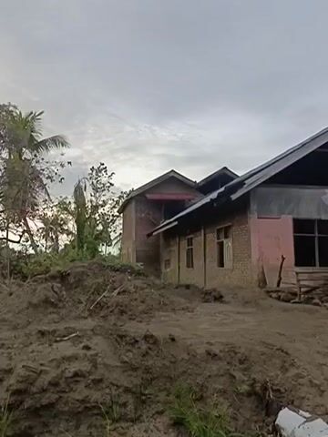 The video shows a semi-finished or partially damaged building. The exterior walls are made of brick and some sections are covered in what appears to be stucco or plaster, with some areas showing exposed brick. The roof is constructed of corrugated metal and wood. There are several windows, some of which are boarded up or have debris in front of them. In the foreground, there is a large pile of dirt or mud, with some vegetation growing in it. A row of sandbags is visible along the bottom of the frame, suggesting efforts to manage water or prevent erosion. In the background, there are palm trees and another small building. The sky is overcast. The overall impression is one of a rural or undeveloped area, possibly affected by natural elements. There are no people visible in the video.