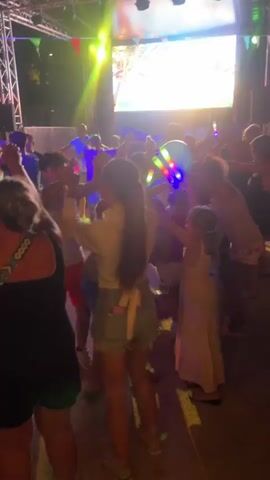 A group of people, primarily children and young adults, are dancing outdoors at night. They are gathered in front of a stage with a large screen displaying colorful visuals. The scene is illuminated by stage lights in various colors, including pink, blue, and yellow, creating a festive atmosphere. Several individuals are holding glowing sticks and a blue balloon, which are also lit up. The people are dressed in casual summer clothing, such as t-shirts, shorts, and dresses. The ground appears to be paved. The overall mood is energetic and celebratory, suggesting an event like a party or a festival.