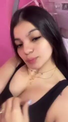 The video shows a young woman with dark hair wearing a black tank top. She is facing the camera with a slight smile. She is wearing a gold necklace and has long eyelashes. Behind her, a pink wall and a small shelf with various items can be seen. An air conditioning unit is visible in the background. She seems to be recording a selfie video.