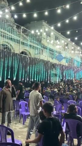 The video shows a crowded outdoor gathering at night, illuminated by numerous string lights and some larger, colored lights. In the foreground, rows of empty purple plastic chairs are visible, suggesting a seating area for an audience. A large crowd of people, primarily men and boys, are gathered in the background. They are standing and milling about, with some seated in chairs that are partially obscured by the crowd. The background features a multi-story building decorated with elaborate strings of lights and banners. A large structure with illuminated letters, possibly a stage or screen, is also visible. The overall atmosphere appears to be festive or celebratory.