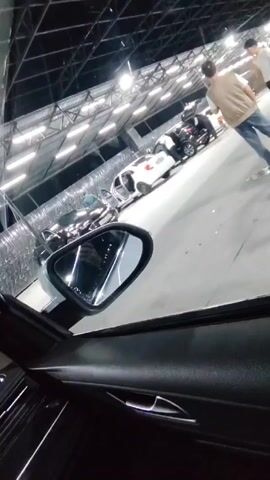 The video shows the interior of a car, possibly at a car wash or detailing center. The dashboard, steering wheel, and side mirror are visible from the driver's seat perspective. Outside, several white cars are parked under bright, industrial-style lighting. People are seen moving around the cars, with some having their trunks open. A sign with an arrow and the word "HIQISH" is visible above the cars. The time displayed on the car's screen is 23:52.