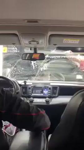Here's a brief description of the video:

The video is shot from inside a car, looking out through the windshield as it goes through a car wash. Soap and water are visible on the windshield, and a brush or cleaning tool can be seen in the driver's periphery. The interior of the car is visible, including the dashboard, seats, and the driver's hands. A charger is plugged into the car's charging port. The central console features a screen showing what appears to be media information.