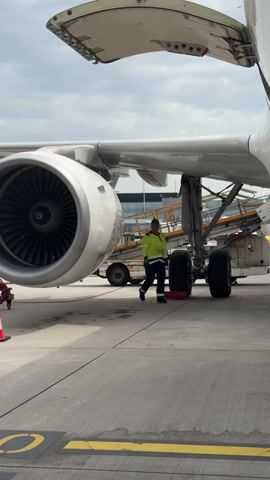 The video shows the wing and engine of a stationary passenger airplane on an airport tarmac. The engine is prominent in the foreground, its large, round housing painted white, and its fan blades are visible inside. The wing extends from the fuselage to the top of the frame, with one of its service panels open. A set of stairs on wheels, marked "Swissport," is positioned underneath the wing near the landing gear, which is exposed. An airport worker wearing a high-visibility vest is walking towards the rear of the plane near the landing gear. An orange and white traffic cone is visible near the edge of the frame on the left. The tarmac is concrete, with visible sealant lines and painted yellow markings. The sky is overcast.