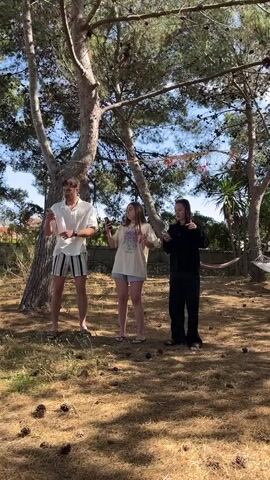 Three young adults, one man and two women, are standing in a yard, each holding a smartphone. They appear to be dancing or moving in sync to music. The man is on the left, wearing a white shirt and striped shorts. The woman in the middle has long hair and is wearing a light-colored shirt and shorts. The woman on the right is wearing a black outfit. The ground is covered in dry grass and pine cones. There are trees scattered around the yard, some with decorations strung between them. A hammock is visible in the background on the right. The sky is visible through the trees.
