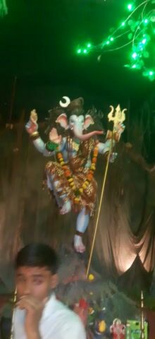 This is a short video clip of a religious idol of the Hindu deity Shiva. The idol is adorned with flowers and jewelry. Shiva is depicted with multiple arms, holding various objects, and with a crescent moon on his forehead. The idol is placed in front of a backdrop of what appears to be a banyan tree, with its aerial roots hanging down. The scene is illuminated by numerous green and white fairy lights, suggesting a festive or ceremonial occasion, possibly at night. The overall atmosphere appears to be spiritual and celebratory. The video is shot in a handheld style, with some blur and motion, giving it a dynamic feel. There is no visible text in the video. The location is identified as Sarangkheda, India, which is a place known for its annual horse fair and associated religious festivals.
