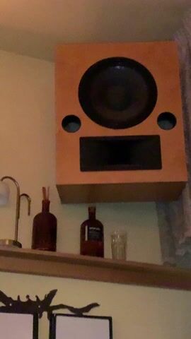A low-angle shot presents a wall-mounted, light brown speaker, beneath which is a shelf displaying a decorative lamp, a dark brown bottle with reeds, another similar bottle, and a clear glass. The speaker has a large, circular black driver, two smaller circular ports, and a rectangular black slot. Sound is emanating from the speaker. The wall is light yellow, and a curtain hangs at the right. A decorative metal piece is partially visible below the shelf.