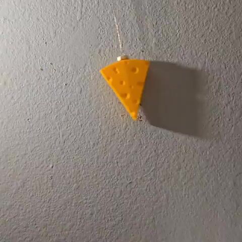 A video shows a yellow cheese slice ornament hanging in front of a textured gray wall. The ornament is shaped like a wedge of swiss cheese and has several round holes. The top of the cheese wedge is attached to a silver ornament cap and hanging by a fine silver chain. The ornament swings gently, casting a shadow on the wall.