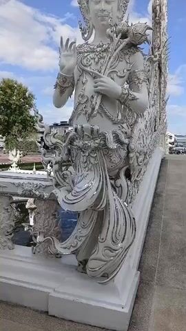 A close-up shot of a white ornate statue of a figure with a headdress and flowing robes, holding a lotus flower in its left hand. The statue is highly detailed with intricate patterns and what appears to be reflective embellishments on the robes. In the background, a cloudy blue sky is visible, with the tops of trees and some buildings. To the right, a segment of the same white ornate structure continues, and a person is partially visible in the distance. The statue stands on a white platform.