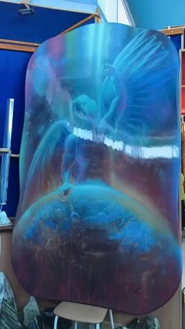The video shows a display of art pieces in a room with blue walls. The first piece is a large painting of an angel standing over the earth. The video then pans to the left, showing other paintings, each mounted on a blue wall. Below the paintings are several rocks and minerals of various sizes and colors, displayed on stands and shelves. The room appears to be a gallery or exhibition space.