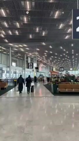 The video shows a view of a large airport terminal. The floor is a shiny gray marble. Overhead, there is an open grid ceiling with multiple bright lights. Signs hang from the ceiling, indicating gate numbers 21, 22, and 23. In the distance, there are shops and seating areas with people walking around. Two people are walking away from the camera, pulling a suitcase. A man walks in the opposite direction, carrying a small suitcase and a tripod bag.