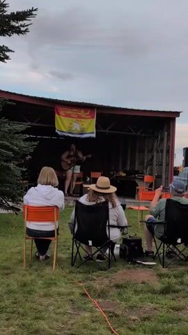 The video shows a person playing a guitar and singing on a small outdoor stage under a cloudy sky. A flag hangs on the back wall of the stage. Three people are seated in chairs watching the performance. The foreground is grass with orange markers on the ground.