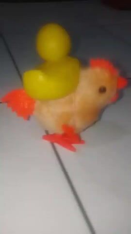 A wind-up toy chicken with a yellow duck on its back walks across a tiled floor. The chicken has orange feet and tail feathers, and its body is brown and fluffy. The duck is bright yellow and has a round head and a beak. The chicken walks from right to left in the frame, with its head bobbing and its feet moving. The tiled floor is white with dark grout lines. In the background, there are some red shoes. The video is slightly blurry and the lighting is dim.