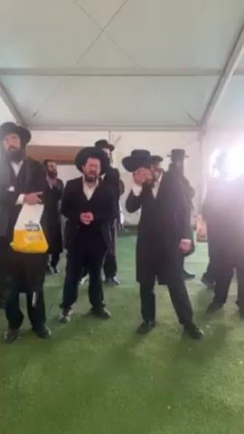 Several men, dressed in traditional Hasidic Jewish attire including black coats, hats, and beards, are gathered under a large, white tent. The tent has a metal frame and white fabric covering. The ground is covered in artificial green turf. Some men are singing, while others stand and watch. One man carries a yellow shopping bag with writing on it. The men appear to be socializing and some are greeting each other. The overall scene suggests a religious gathering or celebration.