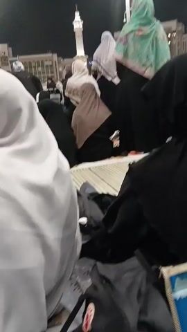 The video shows an outdoor night scene of a crowd of people, mainly women, sitting on the ground in what appears to be a public square or courtyard. The majority of the people are wearing hijabs or other head coverings in various colors including black, white, and shades of brown and green. In the background, several tall buildings are visible, one with a minaret. Streetlights illuminate the area. The audio contains the sounds of someone singing or chanting, possibly in Arabic.