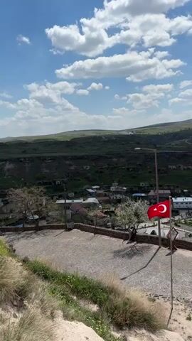 The video presents a panoramic view from a high vantage point overlooking a valley in Acıgöl, Turkey, under a clear blue sky with scattered white clouds. The camera slowly pans from left to right, revealing the landscape.

In the foreground, there is a cobbled path and a small, grassy area. A red Turkish flag, featuring a white crescent moon and star, is prominently displayed on a flagpole in the mid-ground, gently swaying in the breeze. Below the viewpoint, a village with various buildings, some with red roofs, is nestled among green fields and trees. A white bus is parked on a road below, and a few people can be seen walking on the cobbled path and in the distance. The valley floor is characterized by terraced green fields and some barren areas, leading up to distant rolling hills. The overall impression is one of a serene, rural landscape on a bright, sunny day. No discernible text is visible other than the emblem on the flag.