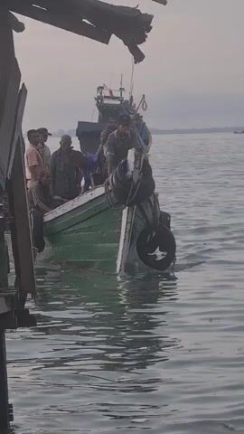 A group of men are gathered on a green wooden boat in the water. The boat appears to be docked or very close to a wooden structure, visible in the foreground on the left. The men are of varying ages and are dressed in casual clothing. Some are standing, others are sitting. The boat has two black tires hanging from its sides, likely used as fenders. In the background, another larger boat is visible on the water, and beyond that, a hazy horizon suggests a distant landmass or the open sea. The water is calm, with gentle ripples. The lighting suggests it is either early morning or late evening. The men appear to be interacting with each other, some looking towards the camera or at other individuals on the boat.