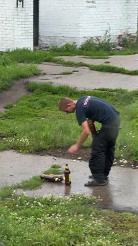 A young man wearing a dark t-shirt and dark pants stands on a wet, asphalt surface. He is bending over and interacting with several beer bottles. In the background, there is a white brick building with a dark doorway and some overgrown grass. The man picks up one of the bottles and places it next to two other bottles that are already on the ground. One of the bottles on the ground appears to be lying on its side. The man then picks up another bottle and appears to be examining it. The scene is outdoors, and the ground is wet, suggesting it may have rained recently.