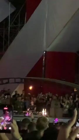 A crowded outdoor music festival is captured from the perspective of someone in the audience. A DJ is visible on a raised platform, silhouetted against bright purple and pink stage lights. The DJ is operating music equipment. The crowd is dense, with many people holding up their phones to record the performance. The atmosphere appears energetic and festive.