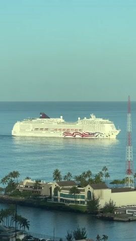 A large cruise ship, painted white with red, white, and blue American flag-inspired graphics, sails across calm, blue ocean water. The ship is seen from a slightly elevated angle, with its port side facing the viewer. It is a sunny day, and the sky is a clear, pale blue, suggesting a pleasant, warm climate.

In the foreground, a lush, green coastline is visible. There are several buildings with white walls and tiled roofs, surrounded by numerous palm trees. The buildings appear to be part of a resort or hotel complex, with some greenery and landscaping. A body of water, possibly a harbor or bay, separates the viewer from the coastline. On the right side of the frame, a tall, red and white lattice tower, likely a radio or communication tower, stands prominently against the sky.

The overall scene evokes a sense of leisure and vacation. The calm water, bright sunshine, and tropical vegetation contribute to a relaxed and inviting atmosphere. The cruise ship itself, a symbol of travel and exploration, is the central focus, poised to embark on or return from a journey. The time of day appears to be morning or late afternoon, given the soft light. No people are distinctly visible on the ship or the shore, emphasizing the grandeur of the ship and the serene landscape. There is no visible text in the image.