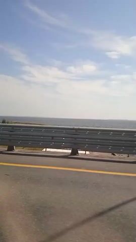 A side-view of a road with a yellow line. A metal guardrail is visible to the left of the road, separating it from a body of water and a line of trees. The sky is blue with some clouds. The footage appears to be taken from a moving vehicle.