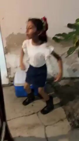 The video shows a girl dressed in a white short-sleeved shirt, a dark blue skirt, and black boots, standing outside in a cracked cement area. She has dark hair pulled back with red flowers in her hair. She's moving her arms and body in a dance-like manner and then walks toward the camera. Behind her is a white wall with peeling paint, a blue and white cooler, and a green leafy plant. A dark wooden object is on the left side of the frame, obscuring the view slightly.