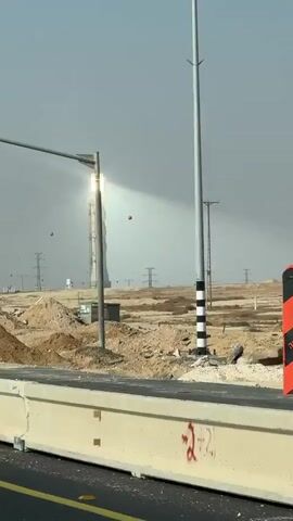 A solar power tower glows brightly in the distance under a hazy sky. In the foreground, a truck drives past a concrete barrier. The landscape appears arid and under construction, with piles of dirt and some temporary fencing. Traffic lights and utility poles are also visible. Red spheres, possibly safety markers or decorations, are scattered around the scene. The video is shot from a moving vehicle.