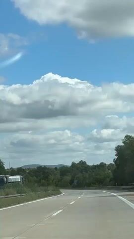 This is a vertical, eye-level shot of a highway in the daytime. The road curves to the right and is lined with white and yellow markings. Lush green trees and bushes grow on either side of the road, with hills in the background. The sky is a bright blue with large, fluffy white clouds. Several vehicles are visible on the highway, including cars, vans, and trucks. There are also road signs with blue backgrounds and white text. The camera appears to be moving, suggesting the footage was taken from a vehicle.