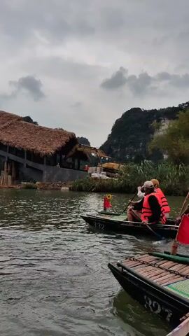 The video shows a river scene with a thatched-roof bridge under construction, with an excavator visible behind it. Several boats are on the water. The people in the boats are wearing life vests. There are forested mountains in the background under a cloudy sky.